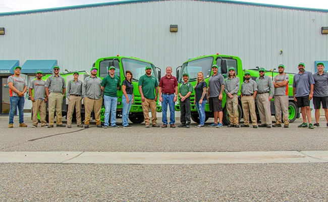 A team photo of Mesa Turf Masters employees and owner with their trucks and office in the background.